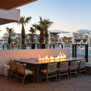 Outdoor dining area with fire pit table and chairs near a waterfront with palm trees and buildings in the background.